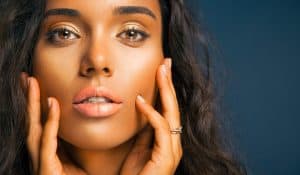 Woman with bronzed skin with hands on face.