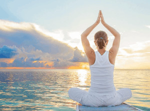 Woman with palms touching above head sitting crossed legged in front of ocean sunset.