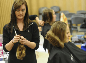 Woman sitting in chair and other woman holding hair. 