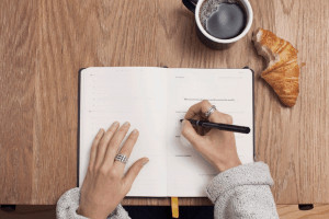 Birds eye view of hands writing in diary next to croissant and coffee mug.