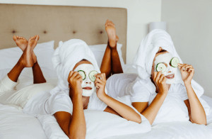 Two girls lying on bed wearing hair towels with face masks and cucumbers on eyes.