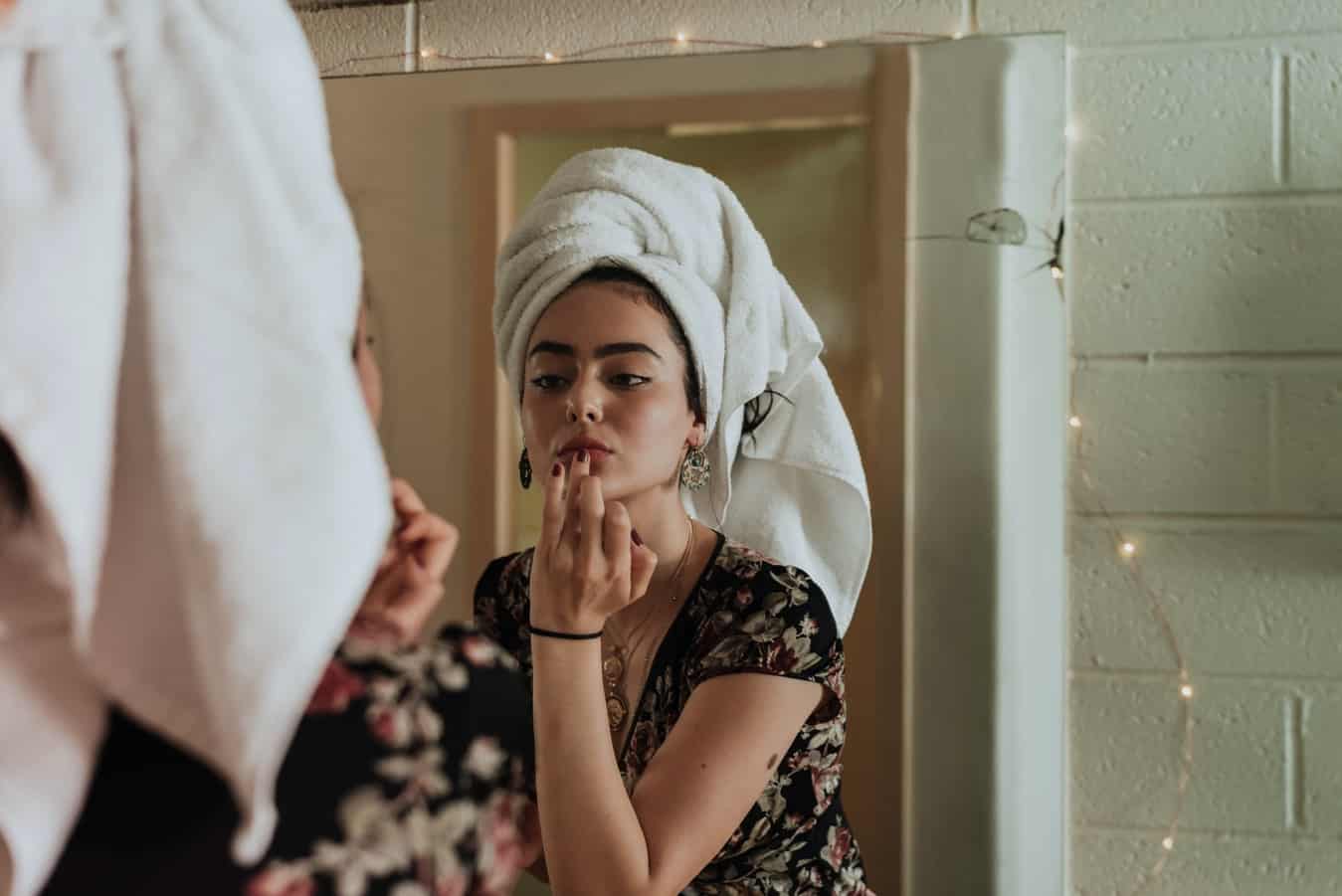 Woman wearing towel on head doing makeup in mirror.